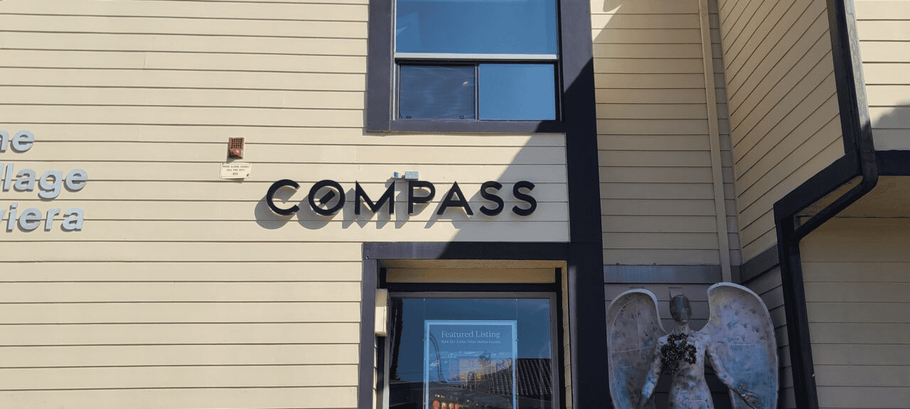 The image depicts a beige building exterior featuring a Custom Signs display reading "Compass" in black letters. Below it, a door with a poster stands near an angel statue holding flowers. Above the door, a window casts shadows on the building, enhancing its timeless charm.