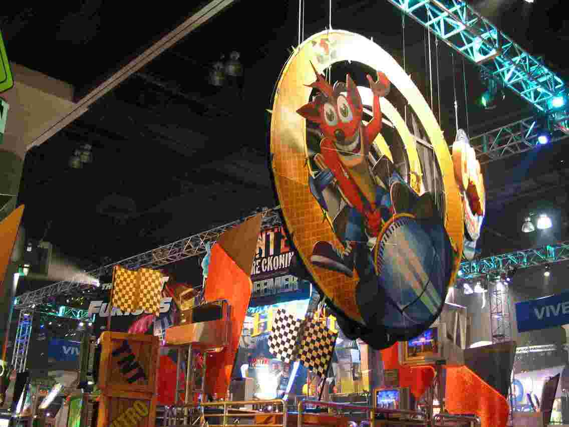 Colorful convention display featuring a large, circular banner with an animated character in a racing vehicle. The background includes checkered flags, hanging lights, and various gaming-related signage. The scene is lively with an energetic atmosphere. 