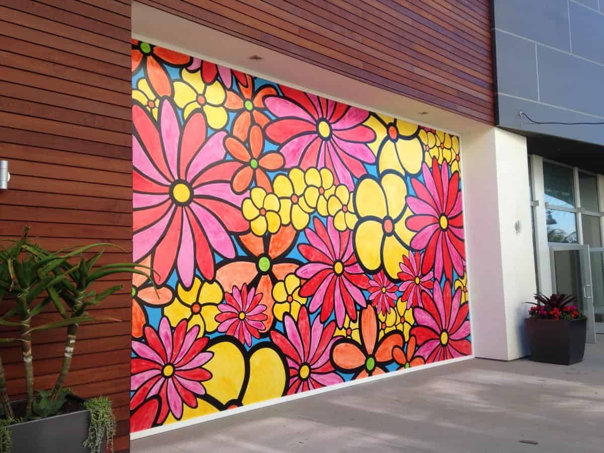 Colorful mural of pink, red, and yellow flowers outlined in black filling a wall space. Set against a wooden and concrete exterior, the vivid floral design contrasts with the neutral tones. A potted plant is placed on either side of the mural, adding to the vibrant aesthetic.