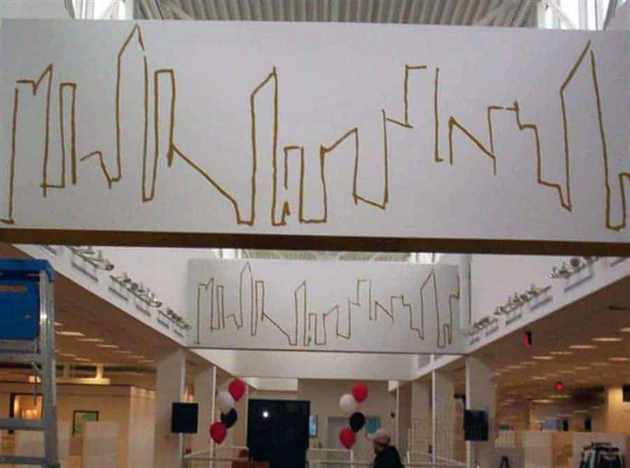 A ceiling installation in a modern building features a series of abstract skyline outlines in gold against a white background. Below, the interior includes columns, red and white balloons, and a person looking up. The space is well-lit, with high ceilings and a contemporary design.