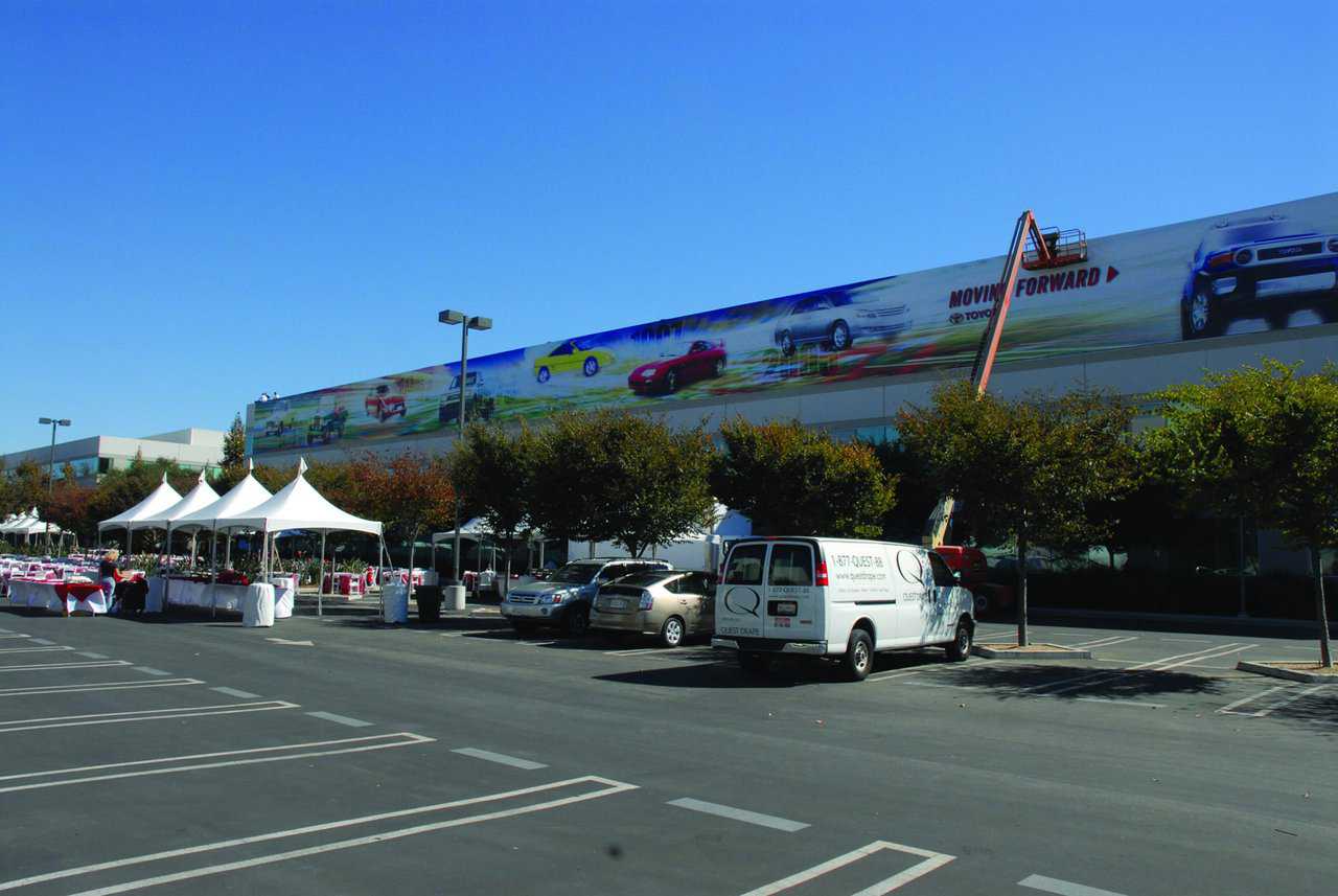 Parking lot with white tents and parked vehicles, including a white van. A large building has a mural featuring colorful cars, with a cherry picker positioned to the right. Trees with reddish-brown leaves line the lot where custom banners flutter gently under the clear blue sky.