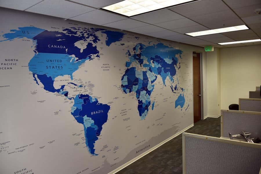 Wall map of the world in an office, featuring countries in shades of blue. The map includes labels for major countries and oceans. The office has gray carpet, beige cubicles, and a brown door on the right with ceiling lighting visible above.