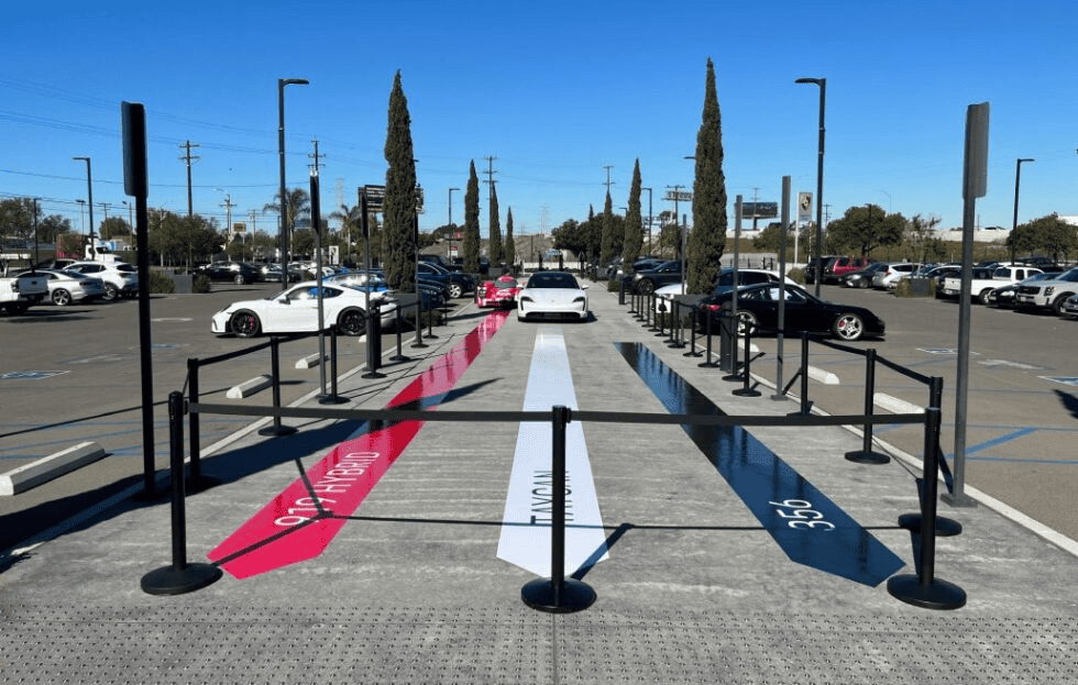 In the car dealership parking lot, new displays feature three sports cars showcased on vibrant floor graphics: a red stripe for "911 Turbo," white for "Taycan," and gray for "356." Tall trees frame the scene against a clear blue sky, while black barriers encircle the gleaming vehicles.
