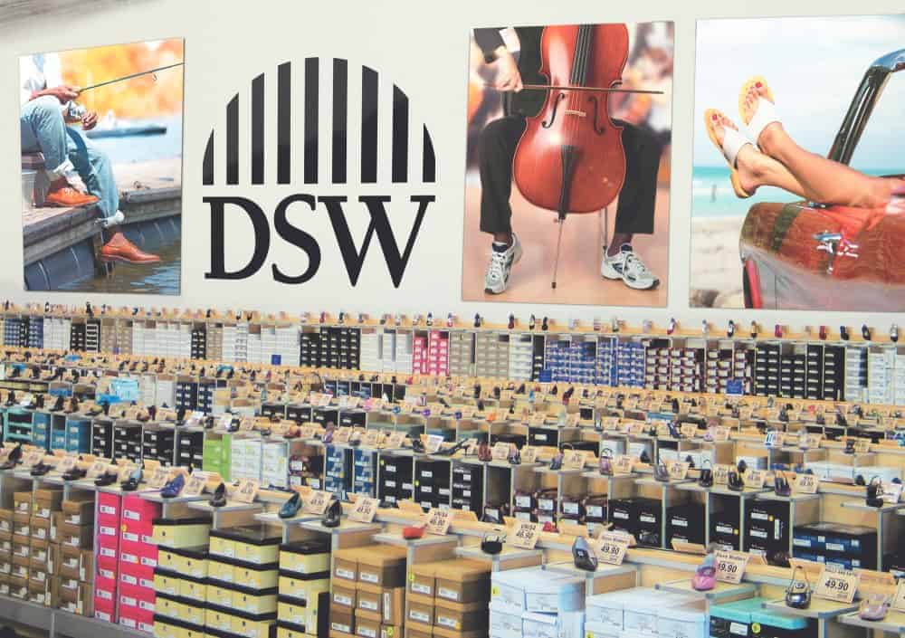 Rows of shoe boxes are neatly stacked on shelves inside a DSW store. Above the shelves, bold retail signage displays the DSW logo, flanked by three photos showing shoes in various settings—by water, playing a cello, and by a car at the beach.