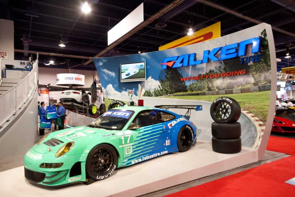 A green Falken Tire race car is displayed at an indoor auto show booth featuring event signage, Falken branding, the slogan “WE GET YOU GOING,” stacked tires, and a small screen above. The brightly lit area has other cars visible in the background. A green Falken Tire race car is displayed at an indoor auto show booth featuring event signage, Falken branding, the slogan “WE GET YOU GOING,” stacked tires, and a small screen above. The brightly lit area has other cars visible in the background.