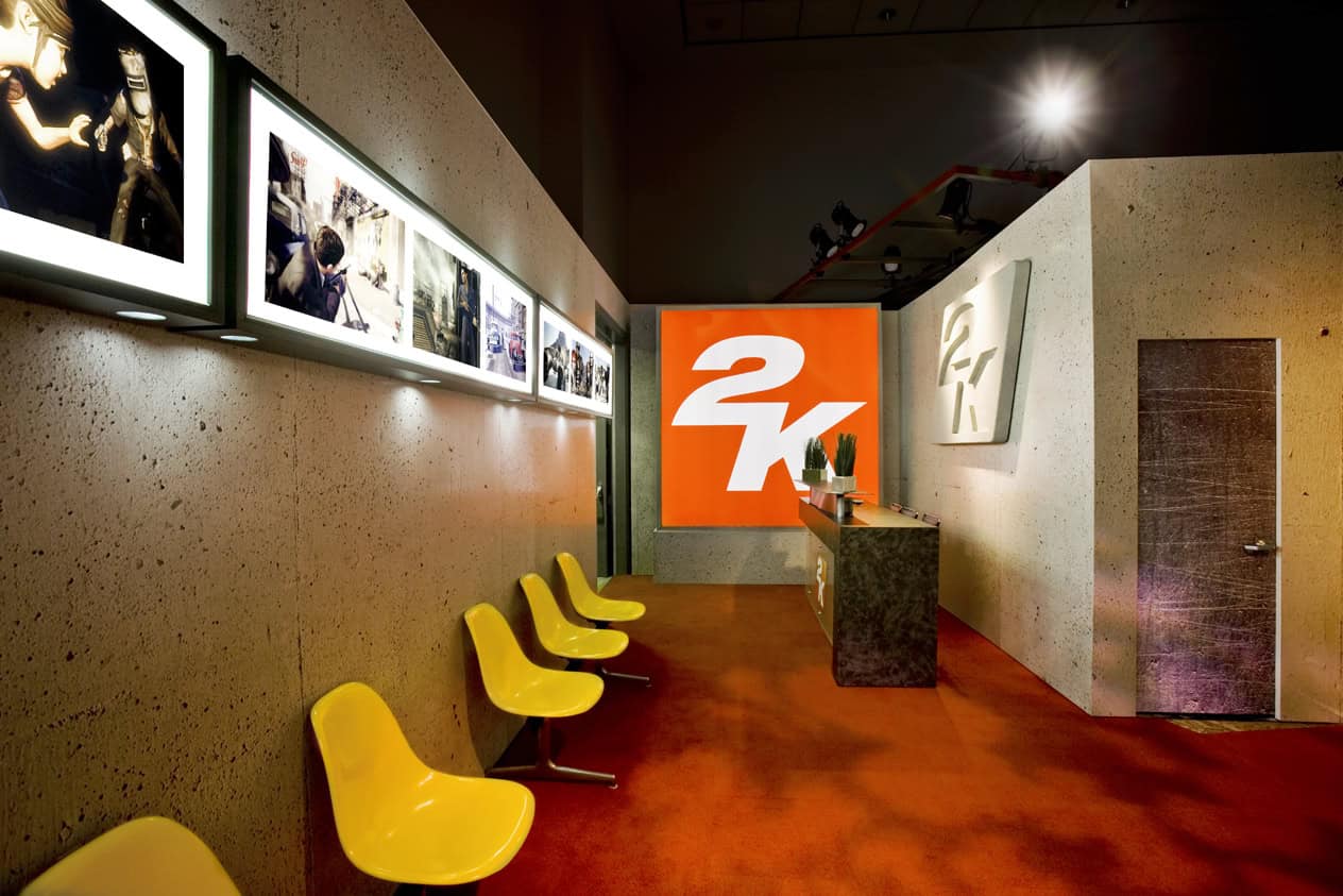 A modern lobby with a row of yellow chairs along a speckled wall displays framed photos and sleek event signage. Opposite, a reception desk stands before a bold orange wall with the white 2K logo. The red floor leads to a metallic door on the right.