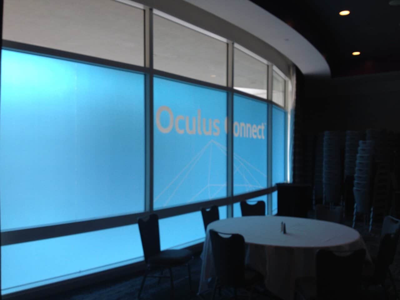 A large blue Event Signage display reads “Oculus Connect” in orange and white text. The room features stacked chairs, a round table with a white tablecloth, and dark flooring, suggesting it’s a conference or event space with bright natural light outside. A large blue Event Signage display reads “Oculus Connect” in orange and white text. The room features stacked chairs, a round table with a white tablecloth, and dark flooring, suggesting it’s a conference or event space with bright natural light outside.