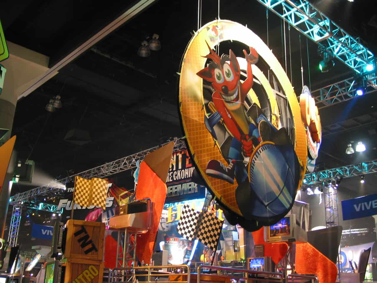 A large circular custom event signage featuring a cartoon fox driving a go-kart hangs above a colorful convention booth adorned with checkered flags, TNT crates, and racing-themed props. Bright lights and other booths are visible in the background.