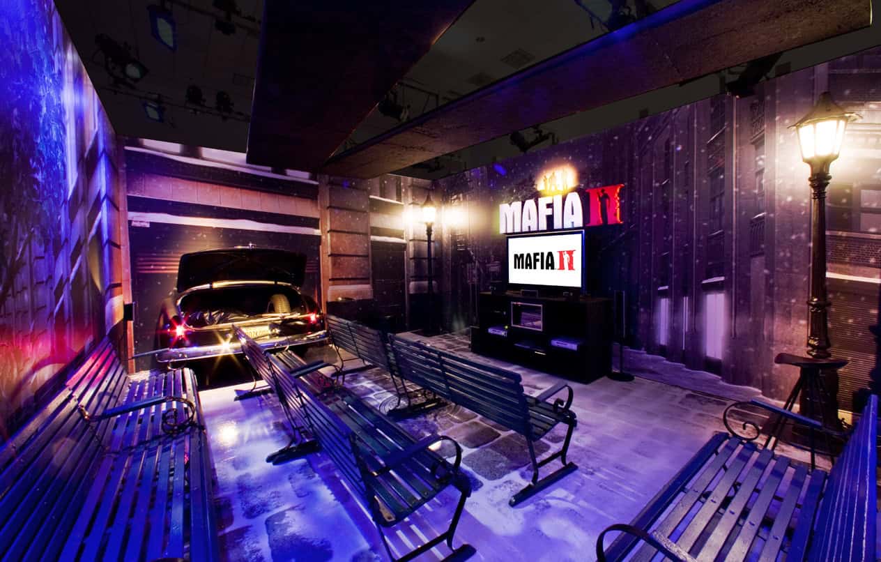 A themed room with streetlights, benches, a vintage car trunk open, and snowy urban backdrop. Event signage and screens display the "Mafia II" logo, evoking a game-inspired night scene reminiscent of 1940s or 1950s city streets.