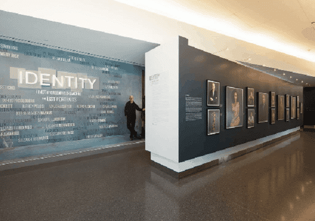 A museum exhibit, reminiscent of a new home page, features the word “IDENTITY” on a blue wall filled with related phrases. A man stands near the entrance, while framed portraits line a dimly lit hallway with polished floors and a white ceiling. 