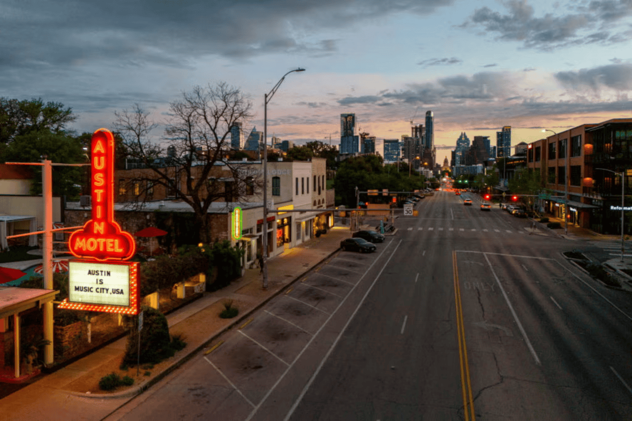 A neon sign reads "Austin Motel" and "Austin is Music City, USA" beside an empty street at dusk in Austin, Texas. Buildings line both sides of the road, with trees and the distant skyline under a cloudy sky in the background.