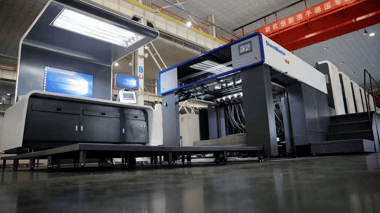 A large, modern industrial printing press for Large Format Printing with a blue and white exterior stands on a polished floor in a Los Angeles factory. Monitors and control panels are visible, while red banners with Chinese text hang overhead.