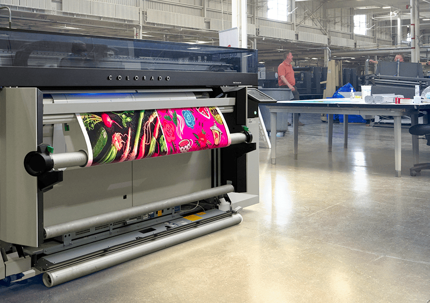 A large industrial printer specializes in Large Format Printing, producing vibrant graphics on a roll of material in a spacious, well-lit San Jose workshop where two people work amid industrial equipment.