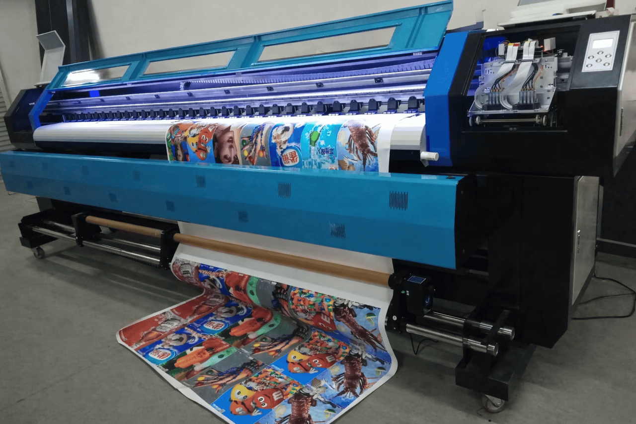A large-format digital printer in Charlotte, North Carolina prints colorful cartoon graphics onto a roll of fabric or vinyl. The blue machine’s control panels glow on the right as vivid, playful images feed out, partially rolled onto the floor.