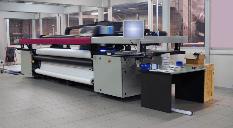 A large format printing setup sits in a clean, bright room with tiled floors and glass walls. Based in Denver, Colorado, it features an industrial printer with rolls of paper, a computer monitor, and a nearby desk holding office supplies and boxes.