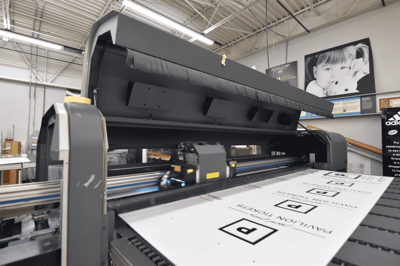 A large-format printer in Houston, Texas prints "Pavilion Tickets" signs in a workshop. The printer head moves across the white paper, while posters and a black-and-white photo of a child hang on the wall beneath exposed ceiling pipes.
