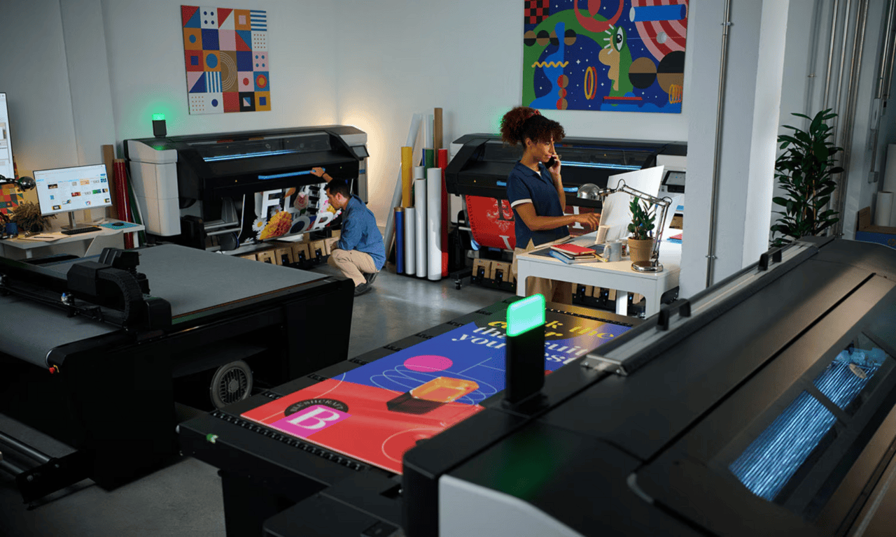In a modern print shop in Memphis, Tennessee, a woman works at a computer desk while another person checks a large-format printer. Colorful posters, rolls of paper, and graphic art decorate the bright workspace with large format printing equipment.