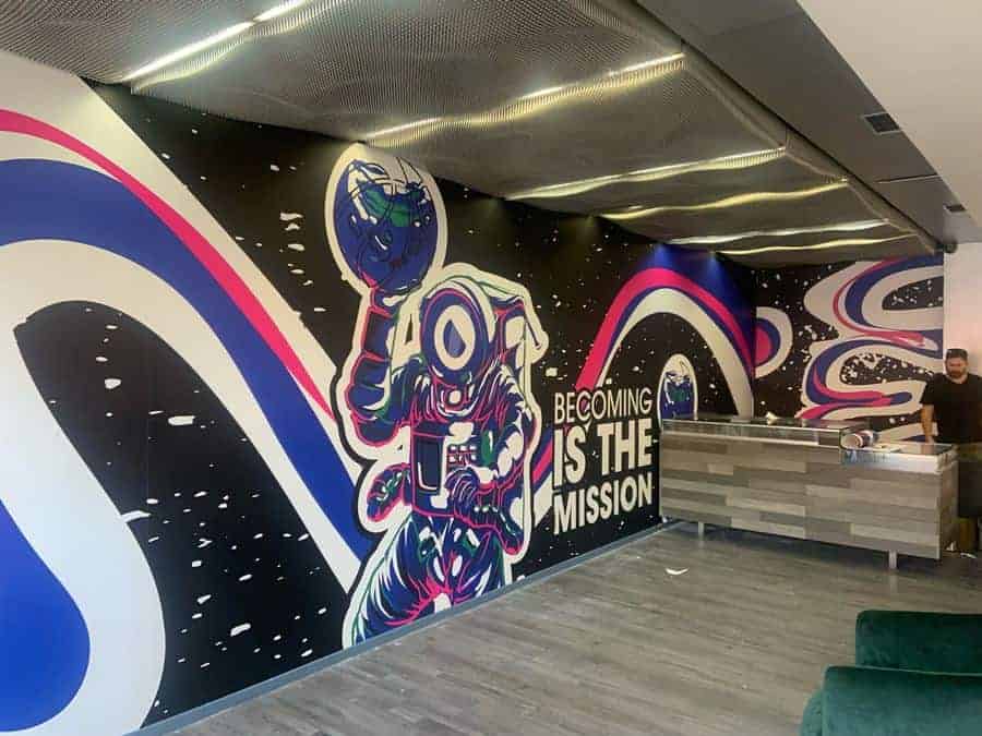 A colorful mural of an astronaut holding a helmet, created with large format printing, covers a wall in a modern New York City room. Swirling blue, pink, and white lines frame the text