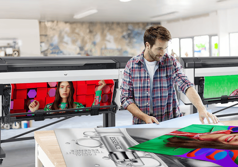 A man wearing a plaid shirt stands in a New York City print shop, working with large printed posters. Two wide-format printers beside him display colorful images, including a woman in front of red cubes and a vibrant green background.