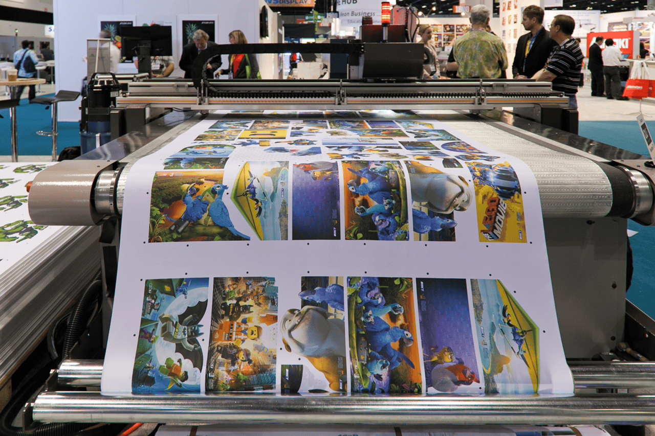 A large format printing setup in Washington DC produces colorful cartoon posters featuring animated characters while several people observe the process at an indoor expo. The vibrant posters are arranged in rows on glossy paper.