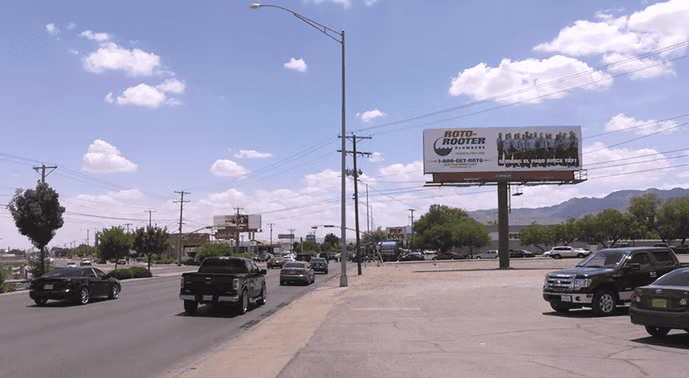 A busy El Paso street with cars driving and parked along the road. Power lines run on both sides. A large Roto-Rooter billboard, created with large format printing, stands to the right; distant mountains rise under a partly cloudy sky.