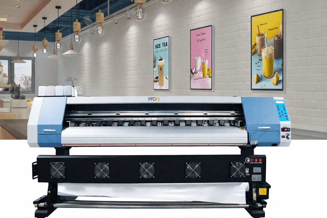 A large-format digital printer with a blue and gray exterior stands in a modern Phoenix cafe. Behind it, framed posters of drinks and desserts decorate a white brick wall. Hanging lightbulbs and seating enhance the airy, contemporary interior.