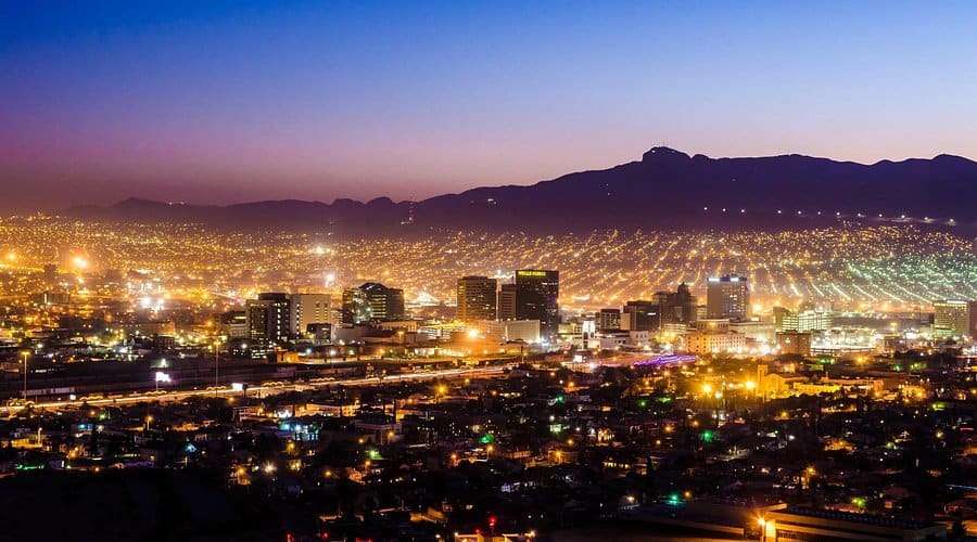 A vibrant Indianapolis cityscape at dusk with brightly lit buildings and streets in the foreground. City lights stretch toward dark mountains under a blue-to-purple gradient sky, perfect for large format printing to capture the lively atmosphere as night falls.