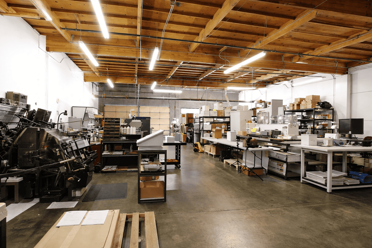 A spacious industrial workshop in Seattle with concrete floors, high wooden ceilings, fluorescent lighting, and organized workstations. Large Format Printing equipment lines the clean, well-lit space alongside shelves of boxes and other machinery.