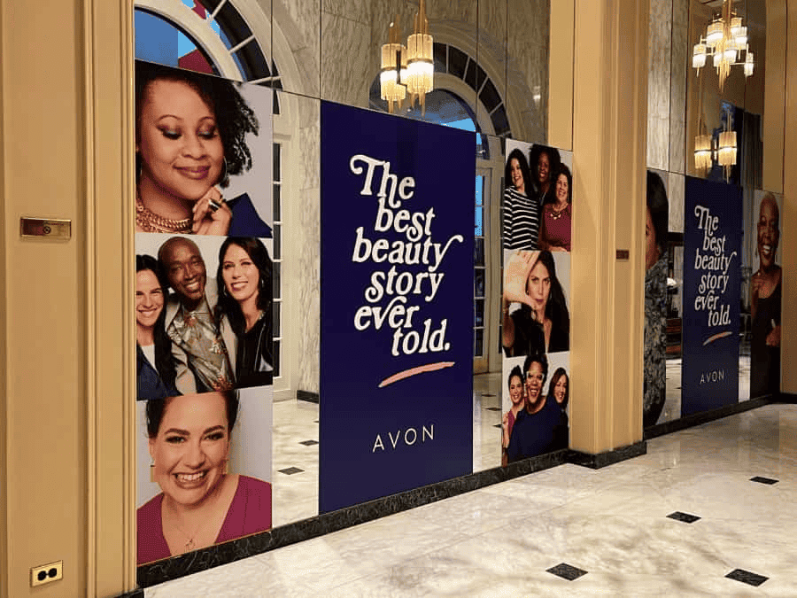 Large posters in a marble-floored Brentwood lobby show diverse smiling people with “The best beauty story ever told. AVON.” Decorative lights hang from the ceiling, and arched windows reflect business printing in an elegant, upscale setting.