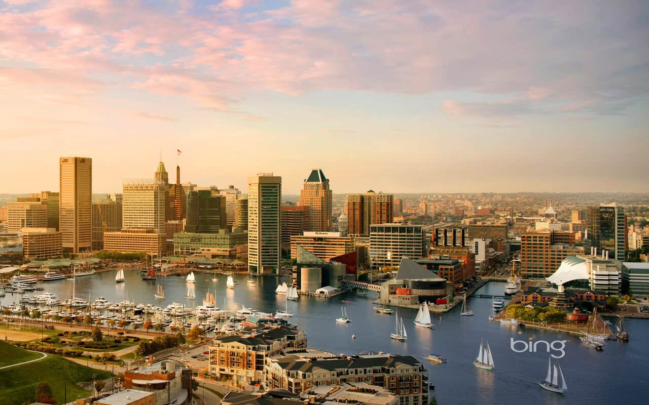 Aerial view of Baltimore’s Inner Harbor at sunset, with sailboats, marina, and a skyline of tall buildings under a pink and orange sky. Landmarks, waterfront attractions, and construction banners are visible; the “bing” logo appears lower right.