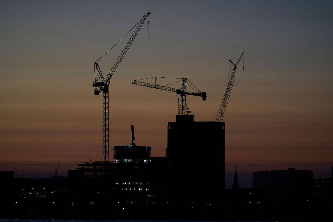 Silhouettes of construction cranes and buildings in Detroit, MI stand against a dusky sky with subtle gradient colors of orange, pink, and purple at sunset. Some windows glow, hinting at ongoing activity as evening falls over the city’s skyline.