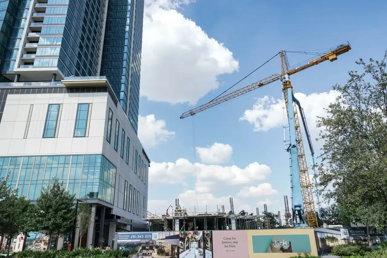 A tall glass building stands next to a Houston, TX construction site with a large yellow crane. Partially-built structures and scaffolding are visible, with construction banners, signs, and green trees in the foreground under a blue sky.
