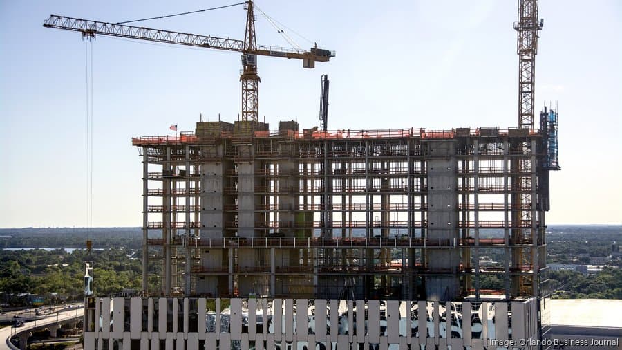 A tall building under construction with exposed concrete floors and steel beams. Two large cranes tower above the structure. Construction materials and scaffolding are visible. In the background, there is a clear sky and distant greenery.