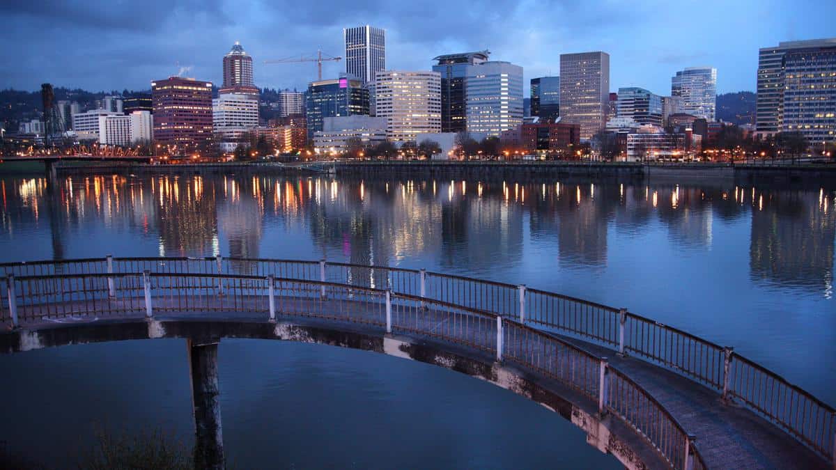 A city skyline at dusk is reflected in a calm river. Tall, modern buildings are lit up against a cloudy blue sky. In the foreground, a curved pedestrian bridge crosses over the water, bordered by railings.