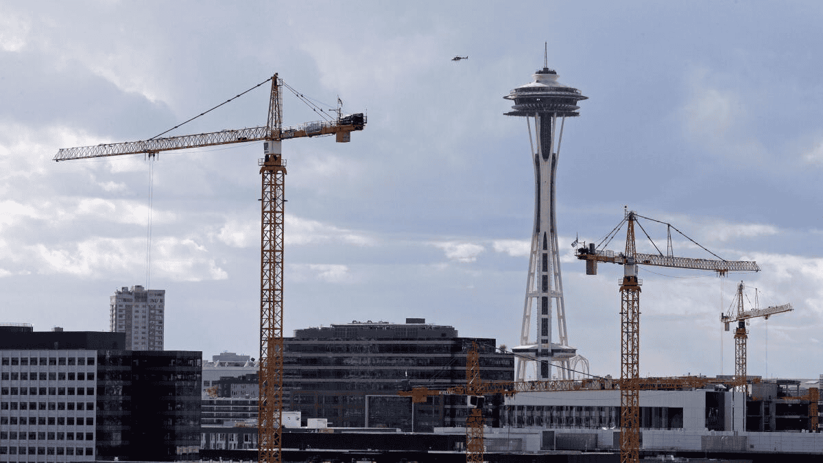 Several yellow construction cranes stand in front of Seattle’s skyline, with the Space Needle prominently visible in the background under a cloudy sky. Mid-rise buildings and a helicopter in the distance complete the urban scene.