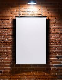 A blank white poster from Layout Posters hangs from two black clips against a red brick wall in Los Angeles, illuminated by a single overhead light that creates a striking spotlight effect.