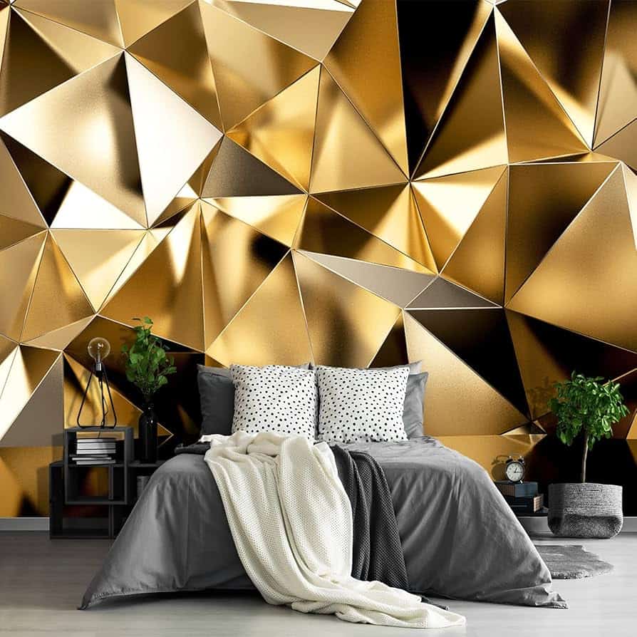 A modern bedroom with a gray bed, polka-dot pillows, and a cream blanket. Two potted plants and a nightstand frame the bed. The wall behind features a bold, gold and black geometric Peel and Stick Wall Murals design with a 3D metallic effect.