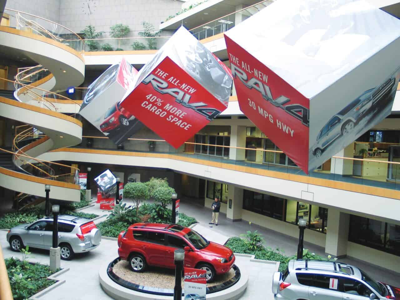 A modern Brentwood atrium with multiple floors features large suspended cubes promoting a red Toyota RAV4 below. The cubes, created by expert business printing, advertise “40% more cargo space” and “31 MPG highway.” Several RAV4s sit among lush indoor plants.
