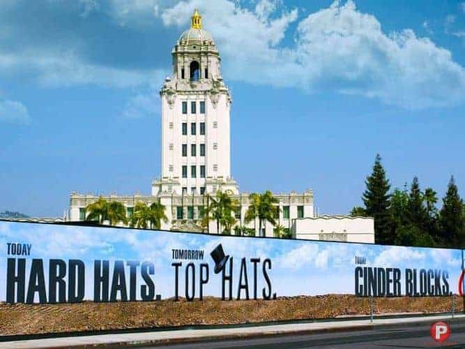 A tall white historic building with a gold dome rises behind a construction site in Brentwood. The blue sky has scattered clouds. A long construction fence displays, "Today HARD HATS. Tomorrow TOP HATS. Then CINDER BLOCKS." Palm trees line the fence.