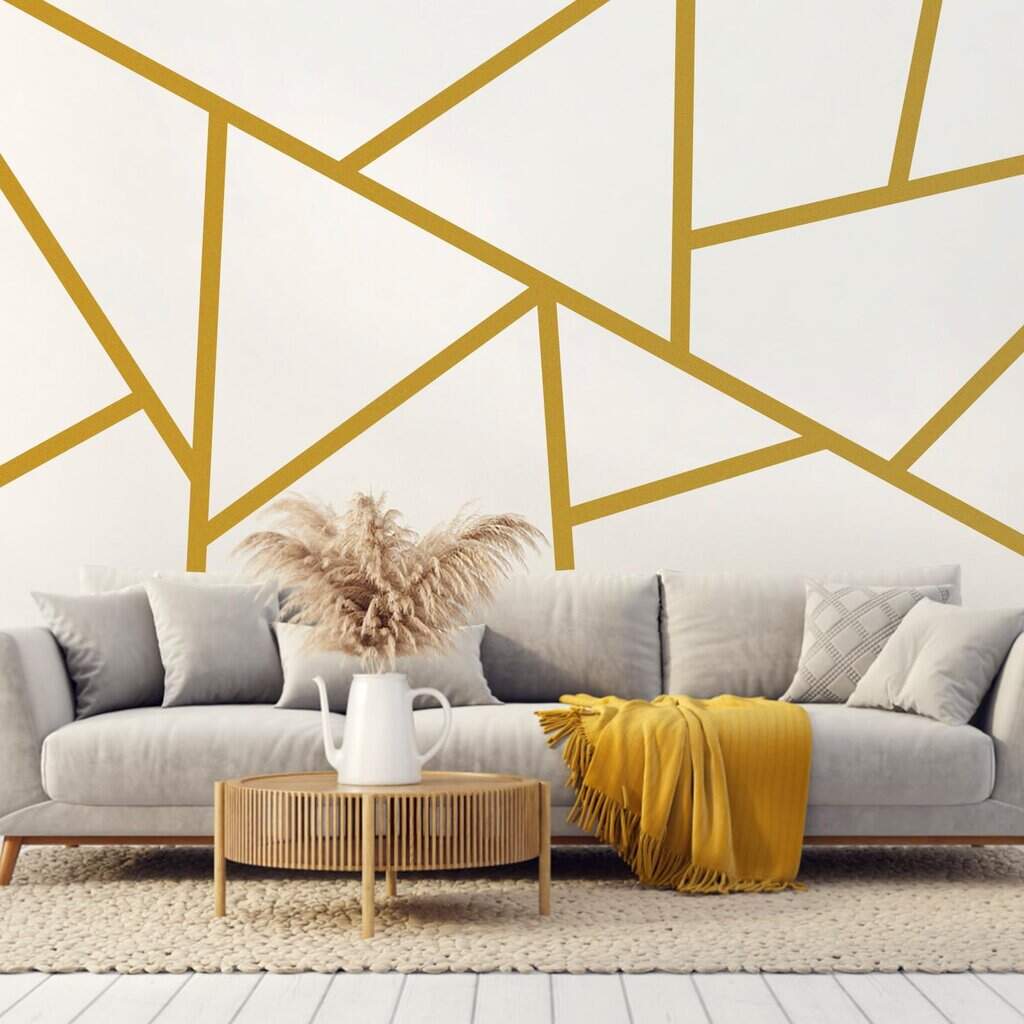 A modern Los Angeles living room with a light gray sofa, yellow throw blanket, and patterned pillow. A round wooden coffee table holds a white vase with dried pampas grass. The wall behind features geometric gold line wall decals on a white background.