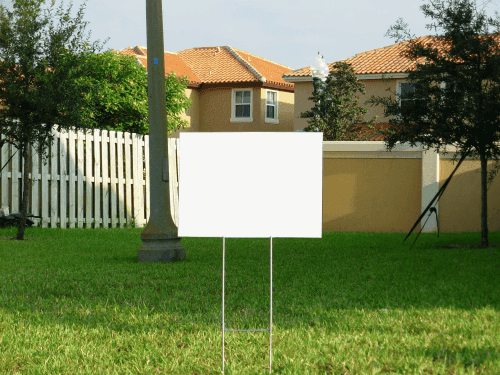 A blank white rectangular yard sign with metal stakes stands on green grass in front of a suburban Los Angeles house with orange roof tiles, a white fence, beige walls, trees, and a utility pole in the background. The sign is empty and unmarked.