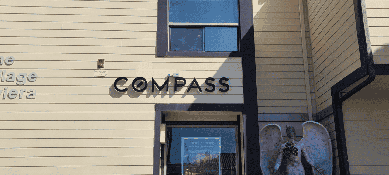 A beige building with black trim features the word "COMPASS" in black capital letters above a glass door. A window sits above the sign, while to the right, all products are watched over by part of a weathered angel statue with wings.