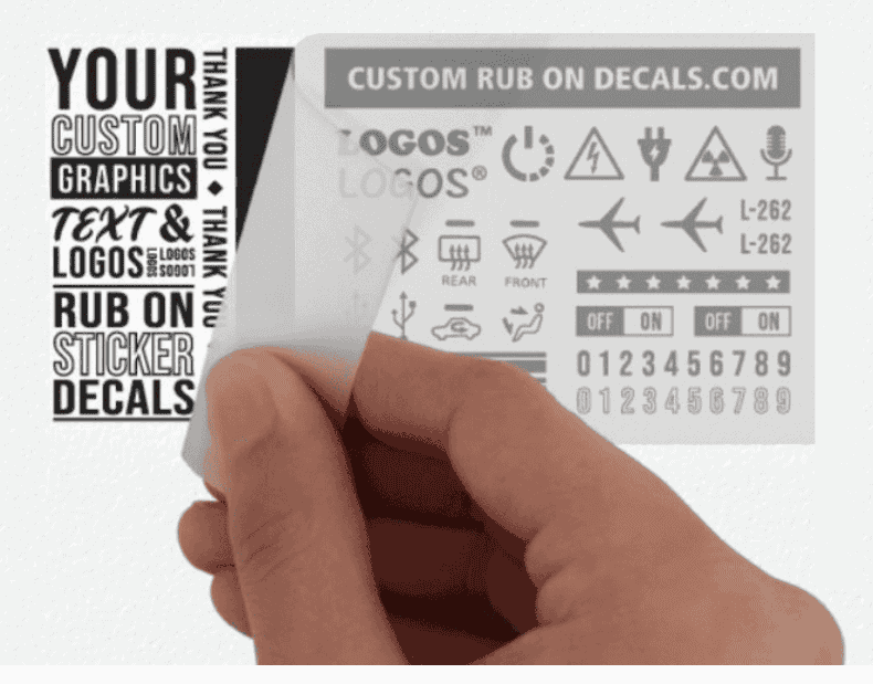 A hand peels a transparent sheet with various black decal designs, symbols, and text from an "All Products" white sheet labeled "YOUR CUSTOM GRAPHICS TEXT & LOGOS RUB ON STICKER DECALS." The top displays icons, letters, numbers, and customizable areas.